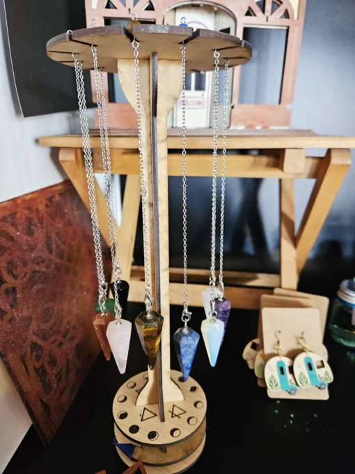 Wooden pendulum display stand with engraved sacred geometry and gemstone pendulums hanging from each slot.