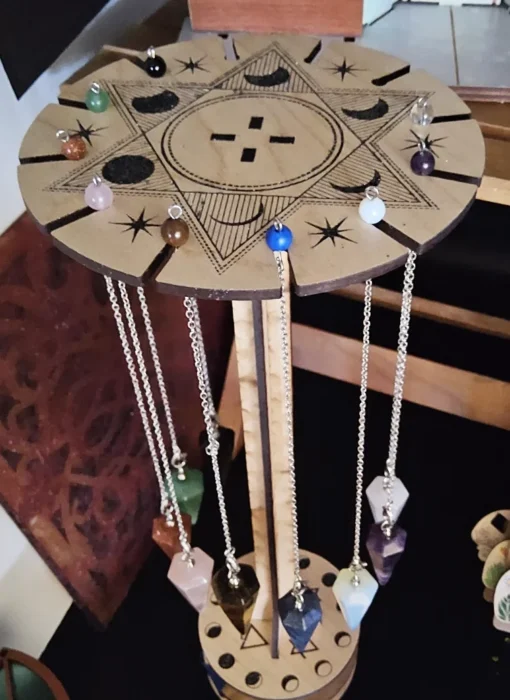 Wooden pendulum holder with multiple crystal pendulums hanging on silver chains, shown on green glitter surface.