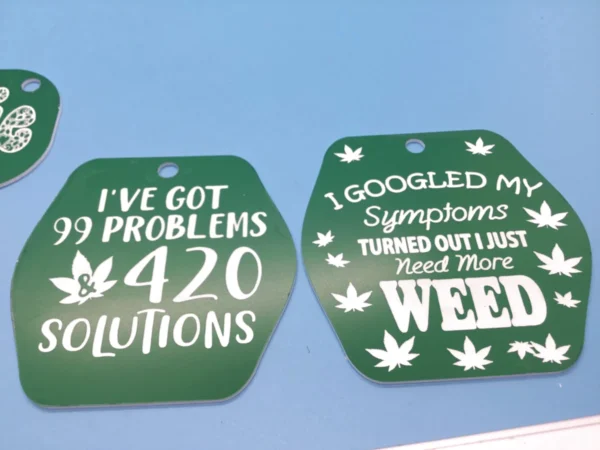 A set of green and white plastic keychains with laser-etched weed quotes, including “99 Problems & 420 Solutions” and “I Just Need More Weed.” These can also be used as magnets.