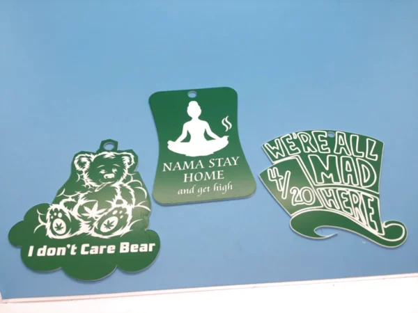 Three green weed-themed keychains featuring humorous designs including “I Don’t Care Bear,” “Nama Stay Home and Get High,” and “We’re All Mad 4/20 Here.”