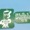 A pair of green acrylic weed keychains—one shaped like a rolled blunt labeled “Kush” and another with the word “Hippie” in a floral mandala font.