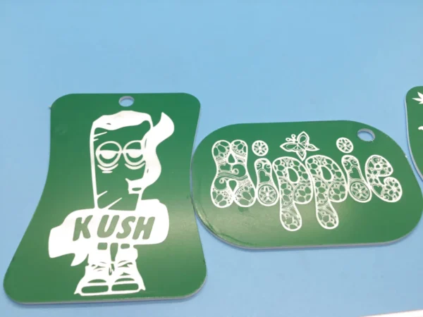 A pair of green acrylic weed keychains—one shaped like a rolled blunt labeled “Kush” and another with the word “Hippie” in a floral mandala font.