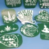 A mixed collection of green acrylic cannabis-themed keychains, laser-etched with stoner sayings and designs like “We’re All Mad 4/20 Here,” “I Don’t Care Bear,” a weed unicorn, lips with a joint, and a meditating figure. A ruler is shown for scale.