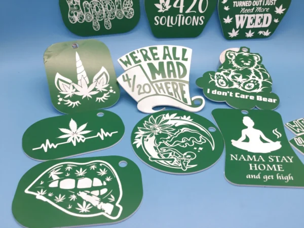 A mixed collection of green acrylic cannabis-themed keychains, laser-etched with stoner sayings and designs like “We’re All Mad 4/20 Here,” “I Don’t Care Bear,” a weed unicorn, lips with a joint, and a meditating figure. A ruler is shown for scale.