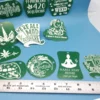 A mixed collection of green acrylic cannabis-themed keychains, laser-etched with stoner sayings and designs like “We’re All Mad 4/20 Here,” “I Don’t Care Bear,” a weed unicorn, lips with a joint, and a meditating figure. A ruler is shown for scale.