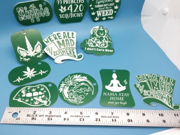A mixed collection of green acrylic cannabis-themed keychains, laser-etched with stoner sayings and designs like “We’re All Mad 4/20 Here,” “I Don’t Care Bear,” a weed unicorn, lips with a joint, and a meditating figure. A ruler is shown for scale.