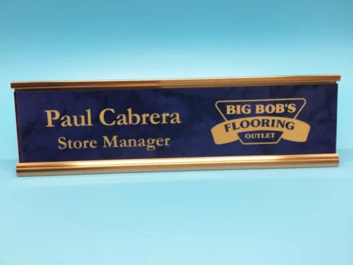 Custom blue and gold engraved name plate with store manager title and business logo, displayed on a bright blue background.