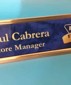 Personalized Custom blue and gold engraved name plate with store manager title and business logo, displayed on a bright blue background.