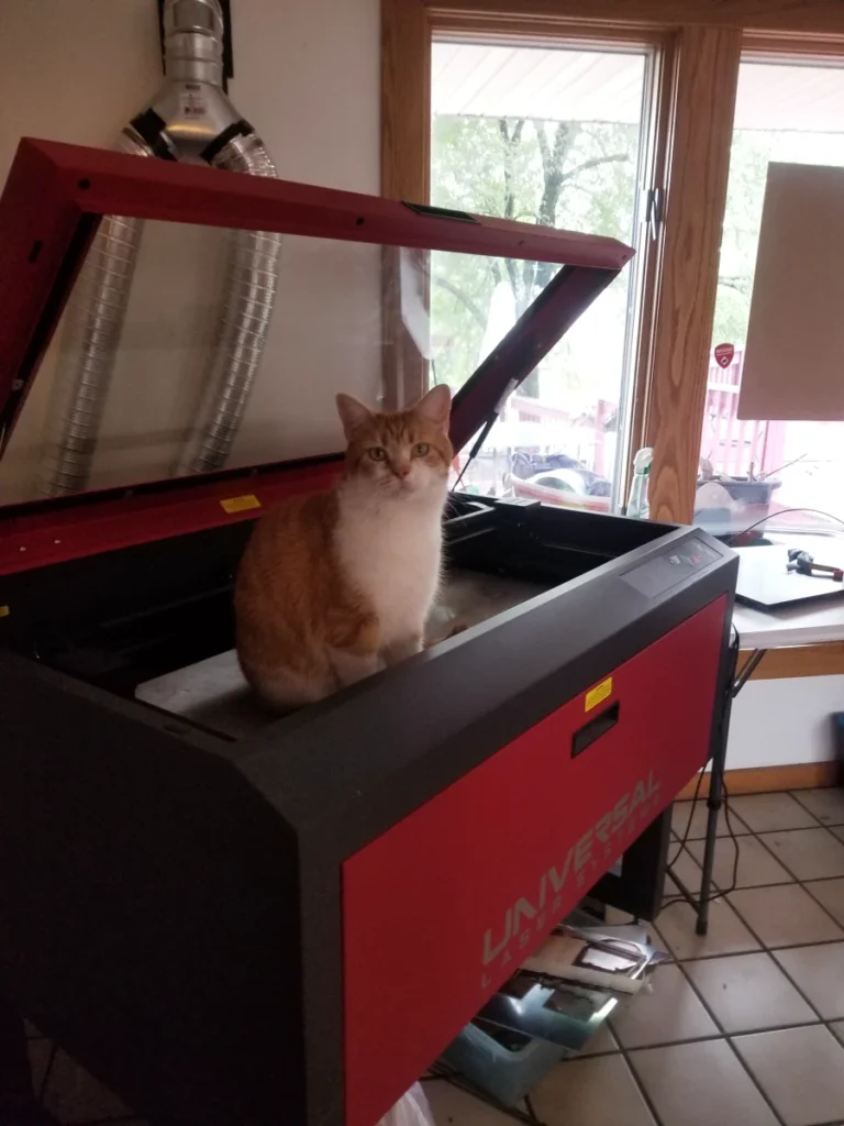 Orange and white cat sitting on a Universal laser engraving machine in a craft workspace.