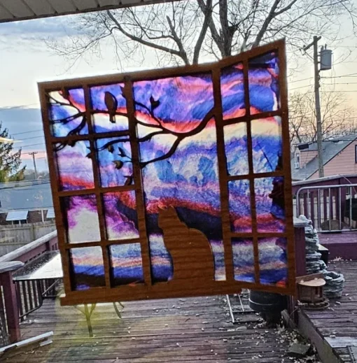 Stained-glass style suncatcher with a silhouette of a cat sitting at a window, gazing at a purple and pink sunset sky with tree branches in the foreground.
